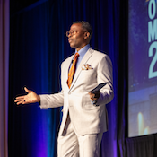 A speaker in a light suit gestures on stage at a conference, engaging the audience under dramatic lighting.