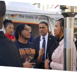 Coolidge student interviews Mayor Muriel Bowser at Holiday Market in front of white tent with two individuals standing beside 